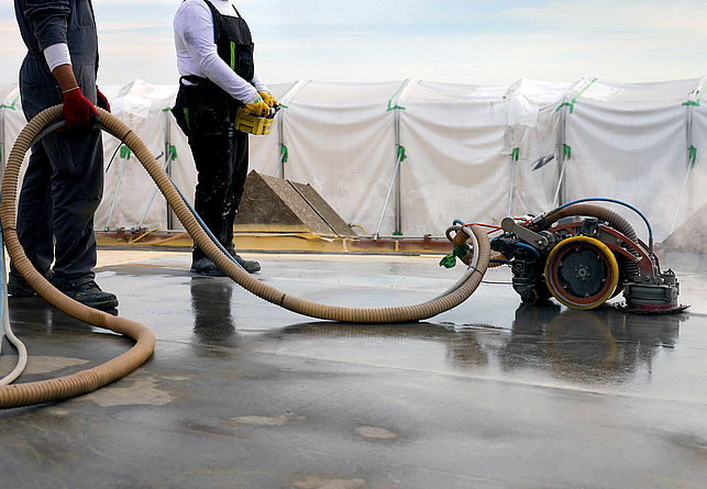 Zwei Personen stehen mit einem Saugroboter auf einer nassen Fläche. Eine Person hält eine Art Fernbedienung in der Hand, die andere hält den Schlauch.