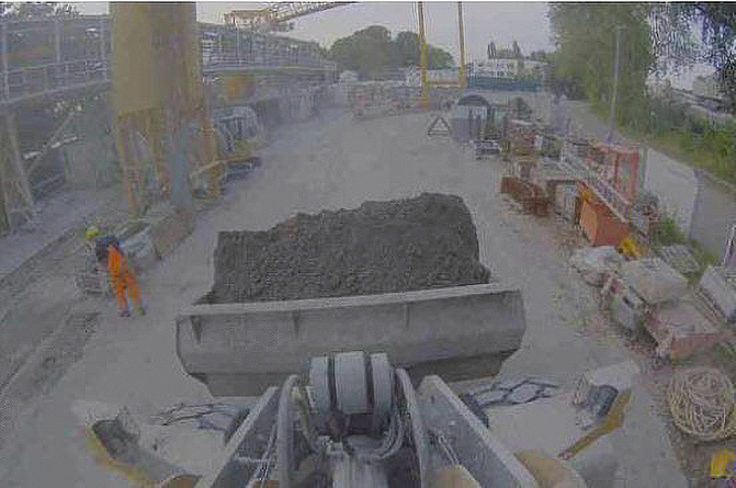 Screenshot des Kamerabildes, welches eine gefüllte Schaufel am Radlader und einen engen Fahrweg mit einer Person am Rand auf einer Baustelle zeigt.