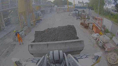 Screenshot des Kamerabildes, welches eine gefüllte Schaufel am Radlader und einen engen Fahrweg mit einer Person am Rand auf einer Baustelle zeigt.