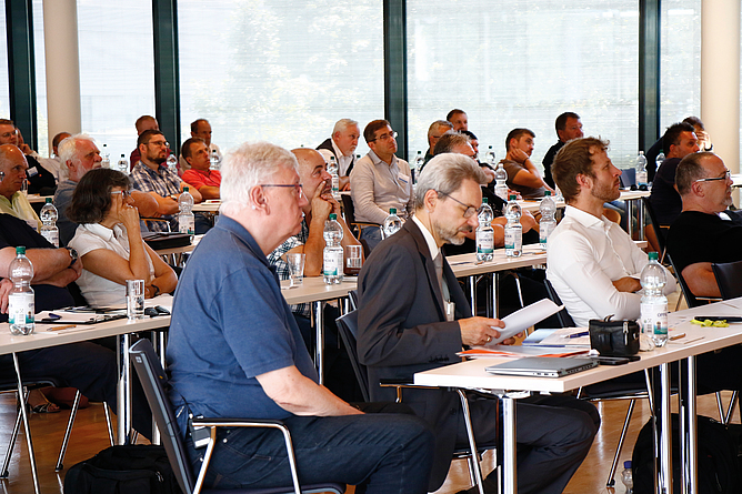 In einem Konferenzraum sitzen zahlreiche Personen an Tischen und hören einer Präsentation zu.