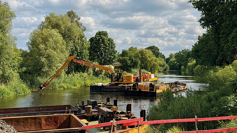 Schwimmplattform mit Bagger auf einem Fluss; im Vordergrund sind Teile einer Brückenbaustelle zu sehen.