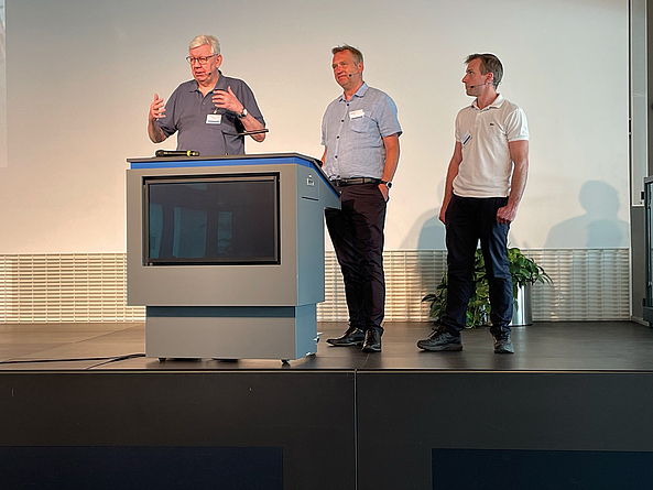 Drei Männer stehen auf einer Bühne hinter einem Rednerpult und sprechen bei einer Veranstaltung.