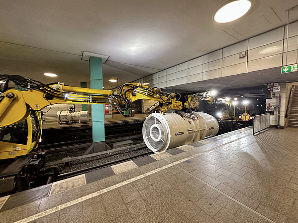 Auf einem unterirdischen Bahnsteig positioniert ein gelber Bagger einen weißen Ventilator im Gleis.