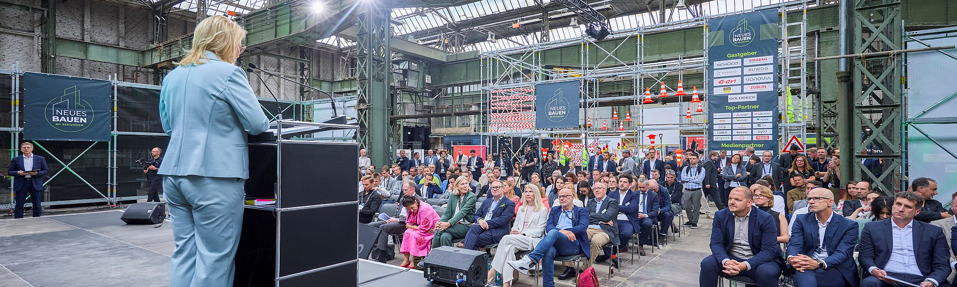 Sabine Poschmann, Parlamentarische Staatssekretärin im Bundesministerium für Wohnen, Stadtentwicklung und Bauwesen Eine Frau steht am Rednerpult auf einer Bühne in einer Industriehalle und spricht zum Publikum.