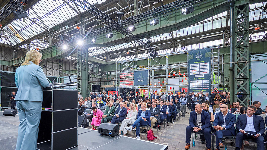 Eine Frau steht am Rednerpult auf einer Bühne in einer Industriehalle und spricht zum Publikum.