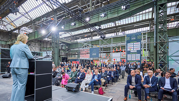 Sabine Poschmann, Parlamentarische Staatssekretärin im Bundesministerium für Wohnen, Stadtentwicklung und Bauwesen Eine Frau steht am Rednerpult auf einer Bühne in einer Industriehalle und spricht zum Publikum.