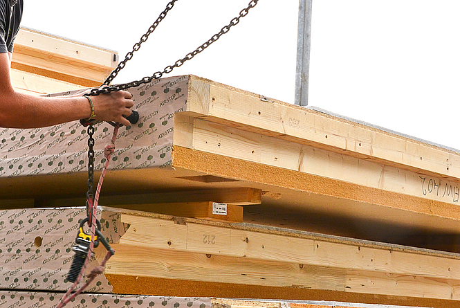 Person, von der nur die Hände zu sehen sind, schraubt eine Halterung, die an einem Stahlseil hängt, in einen Holzblock.