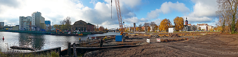 Großflächige Uferbaustelle am Wasser mit Raupenkran, kleinem Container, verschiedenen Materialhaufen und Absperrungen für den Verkehrsweg.