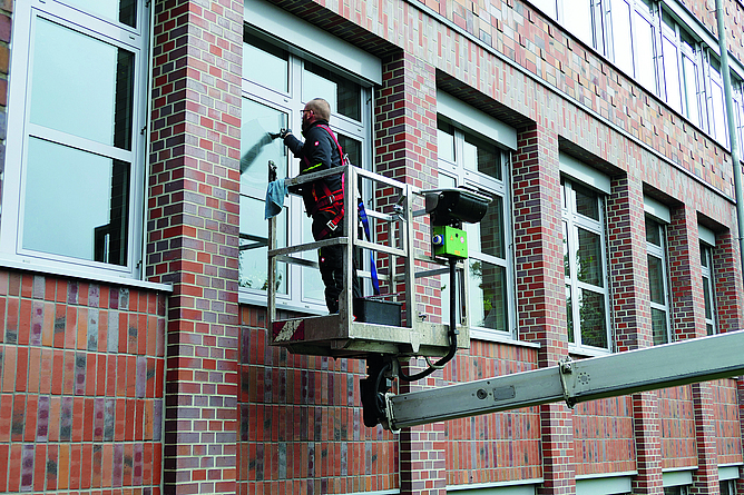Ein Mann, der sich in einer Hubarbeitsbühne befindet, reinigt ein Fenster.Er trägt eine „Persönliche Schutzausrüstung ge­gen Absturz" (PSAgA), Warnkleidung und Helm.