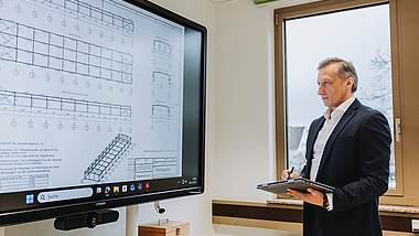 Eine Person im Anzug steht mit einem Tablet vor einem großen Bildschirm und präsentiert eine technische Zeichnung.