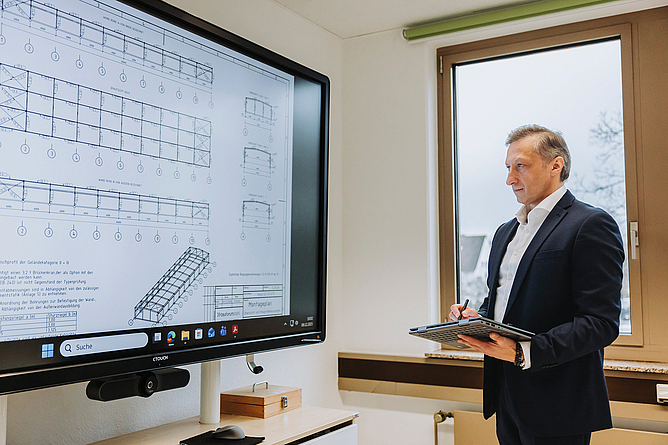 Eine Person im Anzug steht mit einem Tablet vor einem großen Bildschirm und präsentiert eine technische Zeichnung.