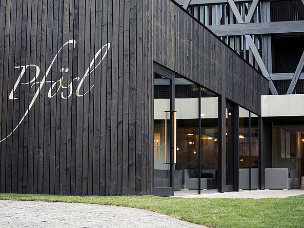Gebäudeteil mit schwarzer Holzverkleidung und dem Schriftzug "Pfösl" sowie großer Fensterfront im Erdgeschoß.