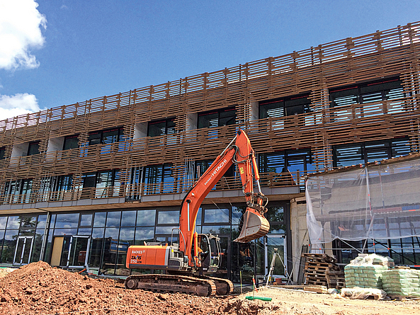 Ein roter Bagger steht vor einem Bauwerk, das zum Teil mit einer Holzfassade versehen ist. Im Vordergrund sind Baumaterialien und eine Kiesgrube zu sehen.