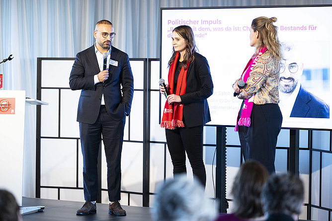 Zwei Frauen und ein Mann mit Mikro auf einem Podium.