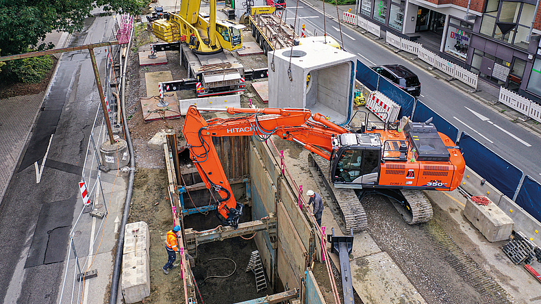 Großbaustelle im Straßenbereich mit einem Kettenbagger, der in einer tiefen Baugrube arbeitet. Im Hintergrund stehen ein Autokran, Betonelemente und abgesperrte Fahrspuren.