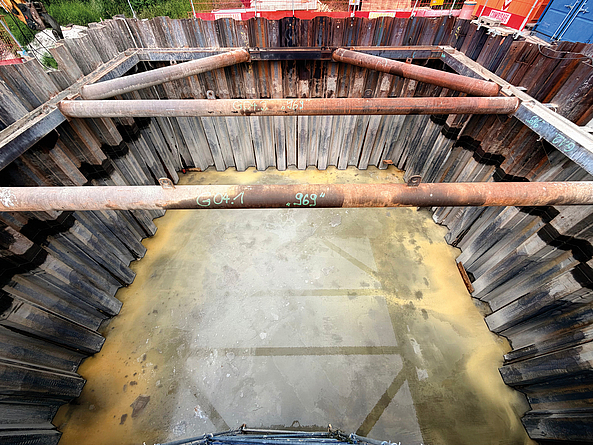 Blick in einen mit Stahlrohren ausgesteiften Spundwandkasten; am Boden der Baugrube steht Wasser.