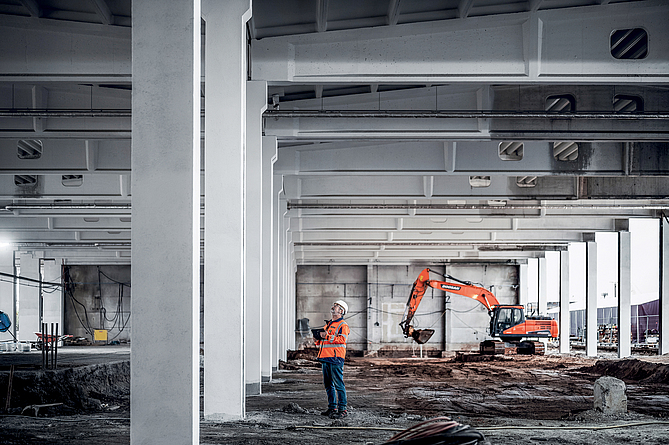 Ein Mann mit Schutzjacke und Helm schaut sich in einer Halle, die gerade saniert wird, die Decke an.
