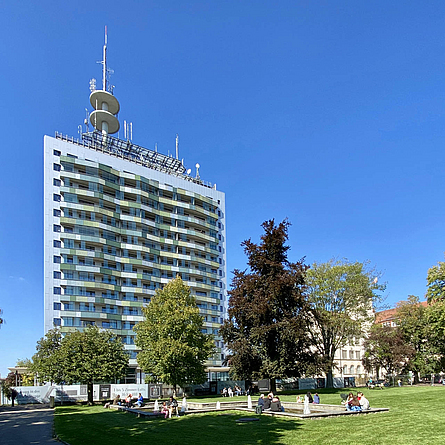 Renoviertes Hochhaus mit bunten Balkonen, davor ist eine Rasenfläche mit Menschen.