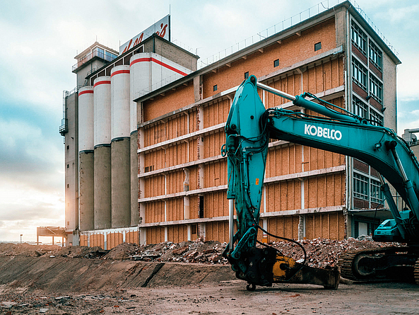 Industriegebäude mit Silos im Rückbau, daneben ein Bagger auf einer Baustelle.
