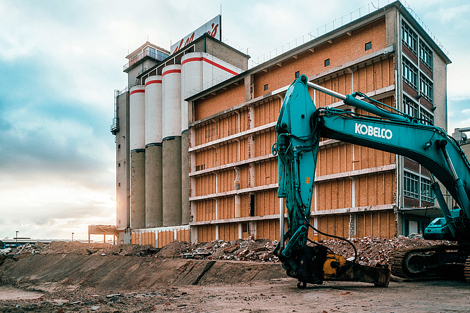 Industriegebäude mit Silos im Rückbau, daneben ein Bagger auf einer Baustelle.