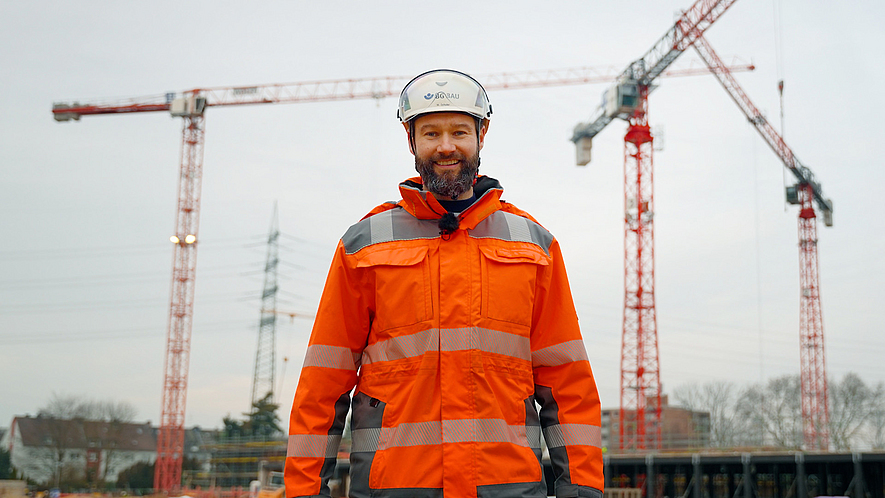 Titelbild des Videos "9 lebenswichtige Regeln im Hochbau": Marc Schuler steht auf einer Baustelle vor drei Kränen.