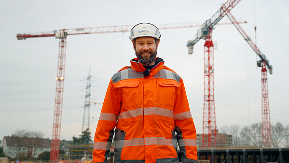Titelbild des Videos "9 lebenswichtige Regeln im Hochbau": Marc Schuler steht auf einer Baustelle vor drei Kränen.