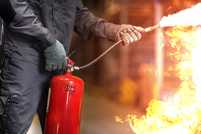 Da PFAS einen Wasserfilm zwischen Brennstoff und Schaum bilden können, werden sie noch oft in Löschschäumen eingesetzt. Um Risiken beim Einsatz zu begegnen, findet eine derzeit eine zunehmende Regulierung von tragbaren Feuerlöschern statt. Ein Mann hält einen Feuerlöscher in der Hand und löscht Flammen.
