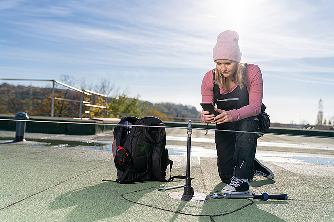 Frau mit Mobiltelefon kniet vor einem Anschlagpunkt mit Seilsystem auf einem Flachdach.