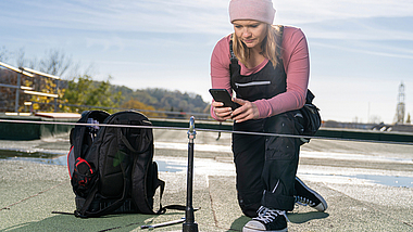 Frau mit Mobiltelefon kniet vor einem Anschlagpunkt mit Seilsystem auf einem Flachdach.
