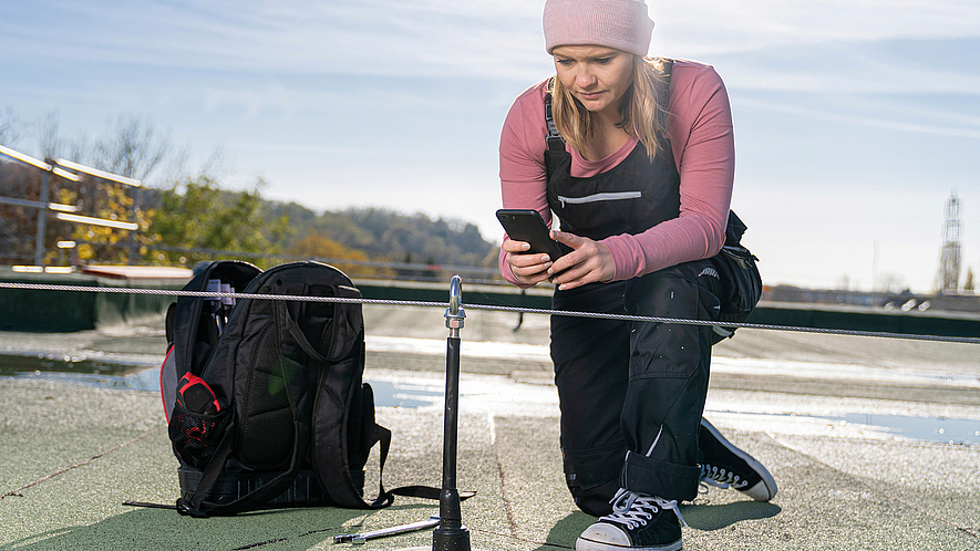 Frau mit Mobiltelefon kniet vor einem Anschlagpunkt mit Seilsystem auf einem Flachdach.