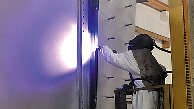 Ein Stahlbauteil wird per von einem Beschäftigten mit persönlicher Schutzausrüstung im Metallspritzverfahren verzinkt.