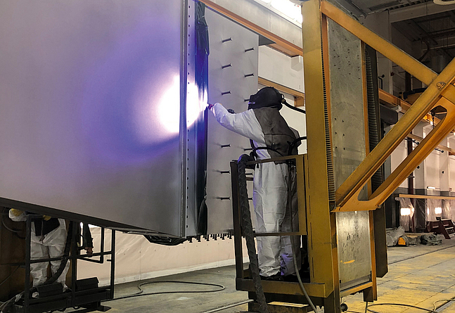 Ein Stahlbauteil wird per von einem Beschäftigten mit persönlicher Schutzausrüstung im Metallspritzverfahren verzinkt.