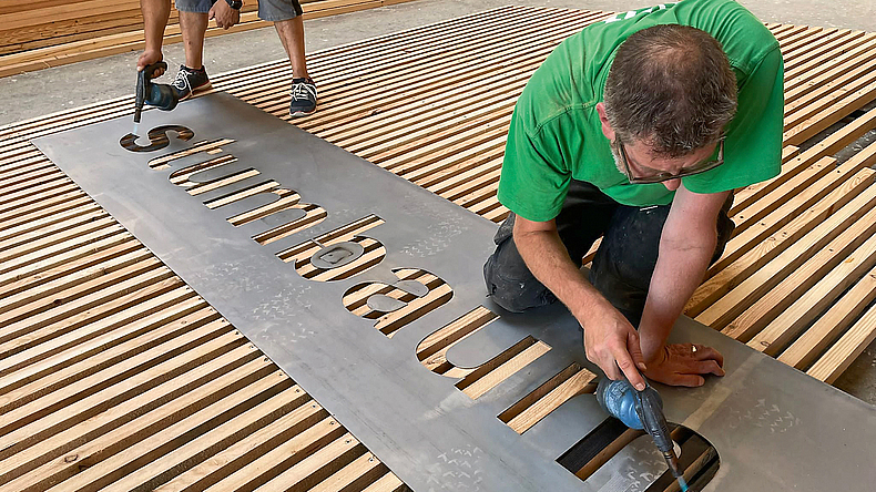 Zwei Männer brennen das Stumbaum-Logo auf das am Boden liegende Holz. Sie verwenden dazu eine Schablone.