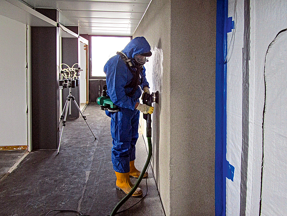 Ein Mann in Schutzanzug und Atemschutzmaske entfernt den Putz von der Wand mit einem Bearbeitungssystem, das mit Absaugung ausgestattet ist.