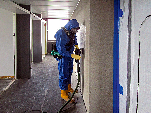 Ein Mann in Schutzanzug und Atemschutzmaske entfernt den Putz von der Wand mit einem Bearbeitungssystem, das mit Absaugung ausgestattet ist.