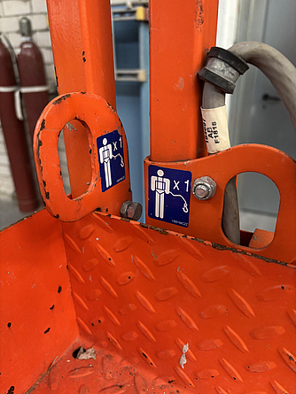 Detailaufnahme einer Arbeitsbühne. Ein blauer Aufkleber weist darauf hin, dass das Tragen von Anseilschutz in jedem Arbeitskorb verpflichtend ist.