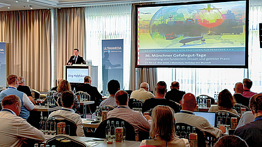 Vortrag vor Publikum in einem Konferenzsaal; auf der Leinwand ist die Folie zu den „Münchner Gefahrgut-Tagen“ zu sehen.