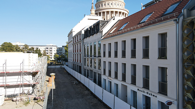 Eine innerstädtische Baustelle verläuft entlang einer sanierten Gebäudezeile mit hellen Fassaden und roten Dächern. Links ist ein eingerüstetes Gebäude zu sehen, im Hintergrund ragt eine Kuppel über die Dächer.