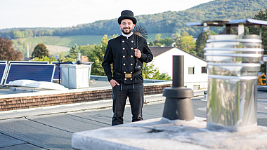 Ein lachender Schornsteinfeger steht auf einem Dach vor einem Schornstein. 