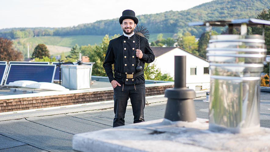 Schornsteinfeger auf dem Dach Ein lachender Schornsteinfeger steht auf einem Dach vor einem Schornstein.