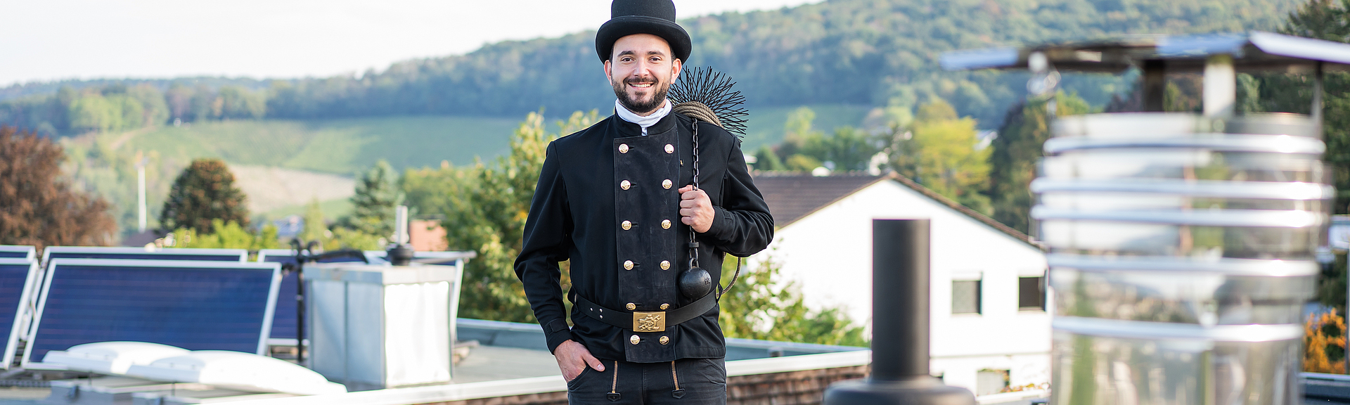 Ein lachender Schornsteinfeger steht auf einem Dach vor einem Schornstein. 