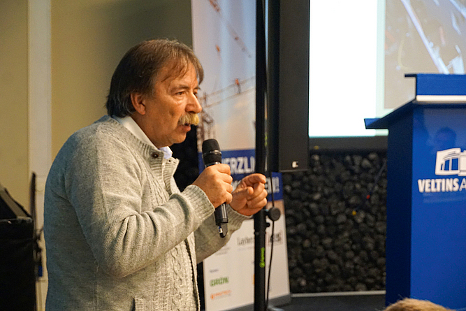 Klaus-Dieter-Wolf spricht mit Mikrofon von der Präsentationsbühne zum Publikum.