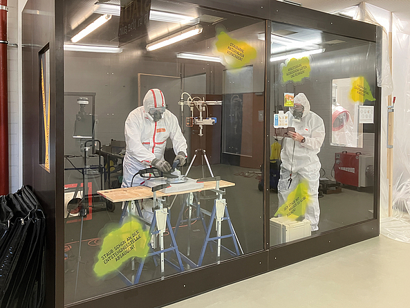 Zwei Personen in Schutzanzügen und Atemmasken messen in einem geschlossenen, gläsernen Raum die Staubbelastung. 