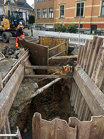 Bauarbeiter schauen in den Graben einer Kanalbaustelle mit Verbauelementen auf einer Straße. Im Hintergrund steht ein Bagger.