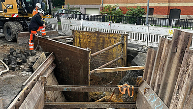 Bauarbeiter schauen in den Graben einer Kanalbaustelle mit Verbauelementen auf einer Straße. Im Hintergrund steht ein Bagger.