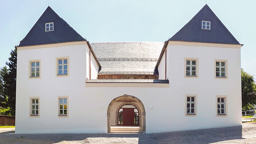 Das „Neue Schloss“ Waldthurn mit dem charakteristischen Rundbogentor nach der denkmalgerechten Sanierung. Weiß saniertes Gebäude mit zwei Flügeln mit Walmdach im Vordergrund. Beide Flügel sind durch einen flachen Bau mit Rundbogentor miteinander verbunden.