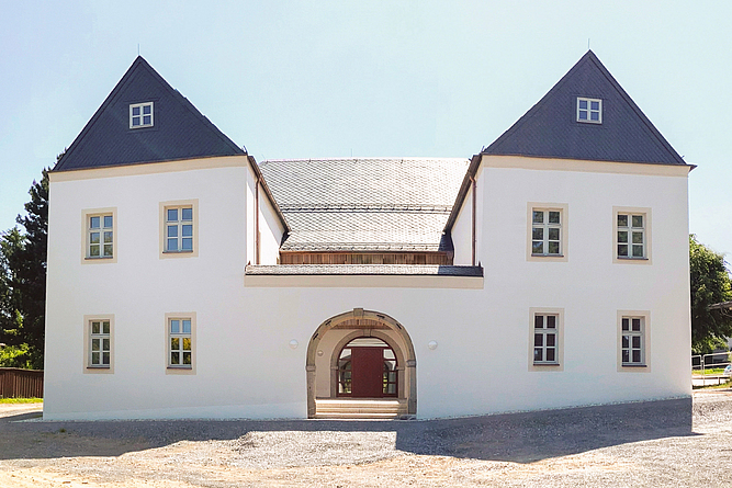 Weiß saniertes Gebäude mit zwei Flügeln mit Walmdach im Vordergrund. Beide Flügel sind durch einen flachen Bau mit Rundbogentor miteinander verbunden.