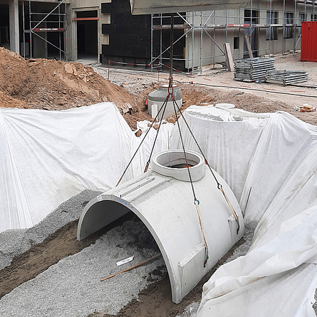 Auf einer Baustelle wird ein vorgefertigtes Stahlbeton-Schachtelement mithilfe eines Krans in eine vorbereitete Baugrube eingesetzt.