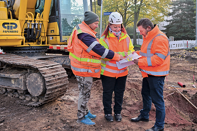 Drei Personen mit Schutzjacken stehen vor einem Baufahrzeug und besprechen einen Plan, den die mittlere Person in der Hand hält.