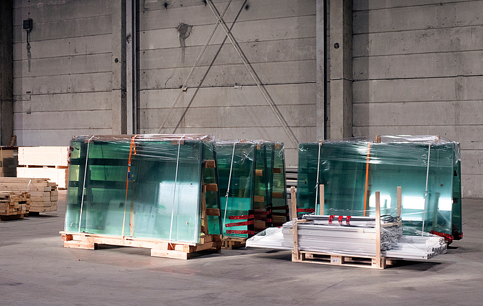 Mehrere große Glasscheiben, verpackt und auf Holzpaletten gelagert, stehen in einer Industriehalle.